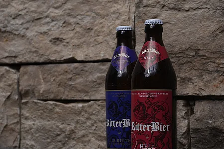 Zwei Bierflaschen mit Etiketten "RitterBier" vor einer Steinwand. Eine Flasche hat ein rotes, die andere ein blaues Etikett.